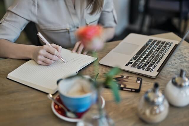 Woman writing in notebook while working at laptop