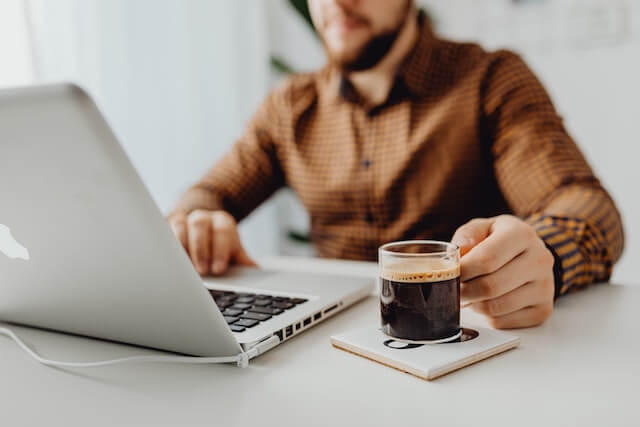 Man sitting at a desk looking at a laptop with clear mug of coffee in hand