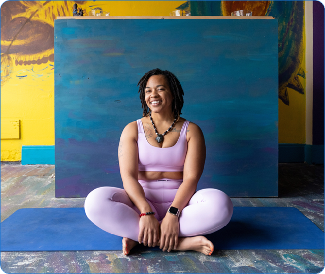 A Black woman sitting cross-legged on a blue yoga mat with artwork behind