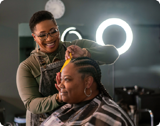 A Black woman braiding another Black woman’s hair with studio lights shining behind