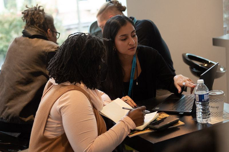 Two people working on a laptop in a room with other people