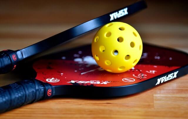 Yellow picklball lying between two black and red paddles on the ground