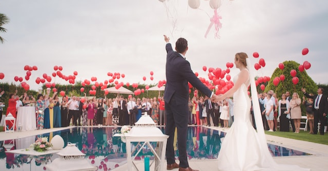 A wedding party holding red balloons, watching the bride and groom as they release white balloons into the air