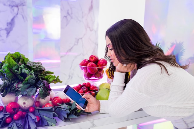 Ayesha Curry Looking into Smartphone with Vegetables on Counter