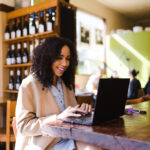 Young woman smiling, sitting at reclaimed wood table while looking at laptop computer working