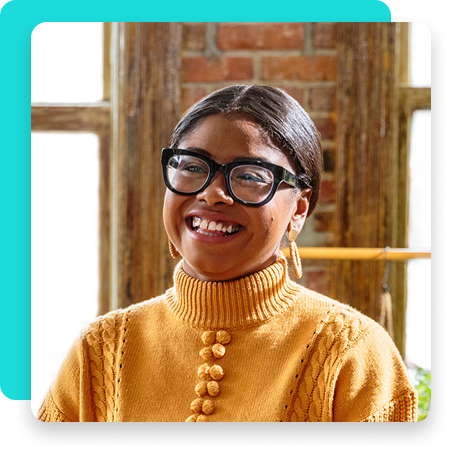 Smiling woman in mustard sweater and glasses in front of brick wall