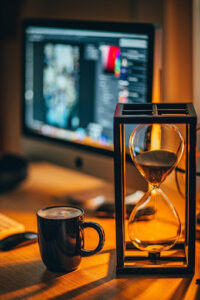 Hourglass sitting next to coffee mug with computer in background