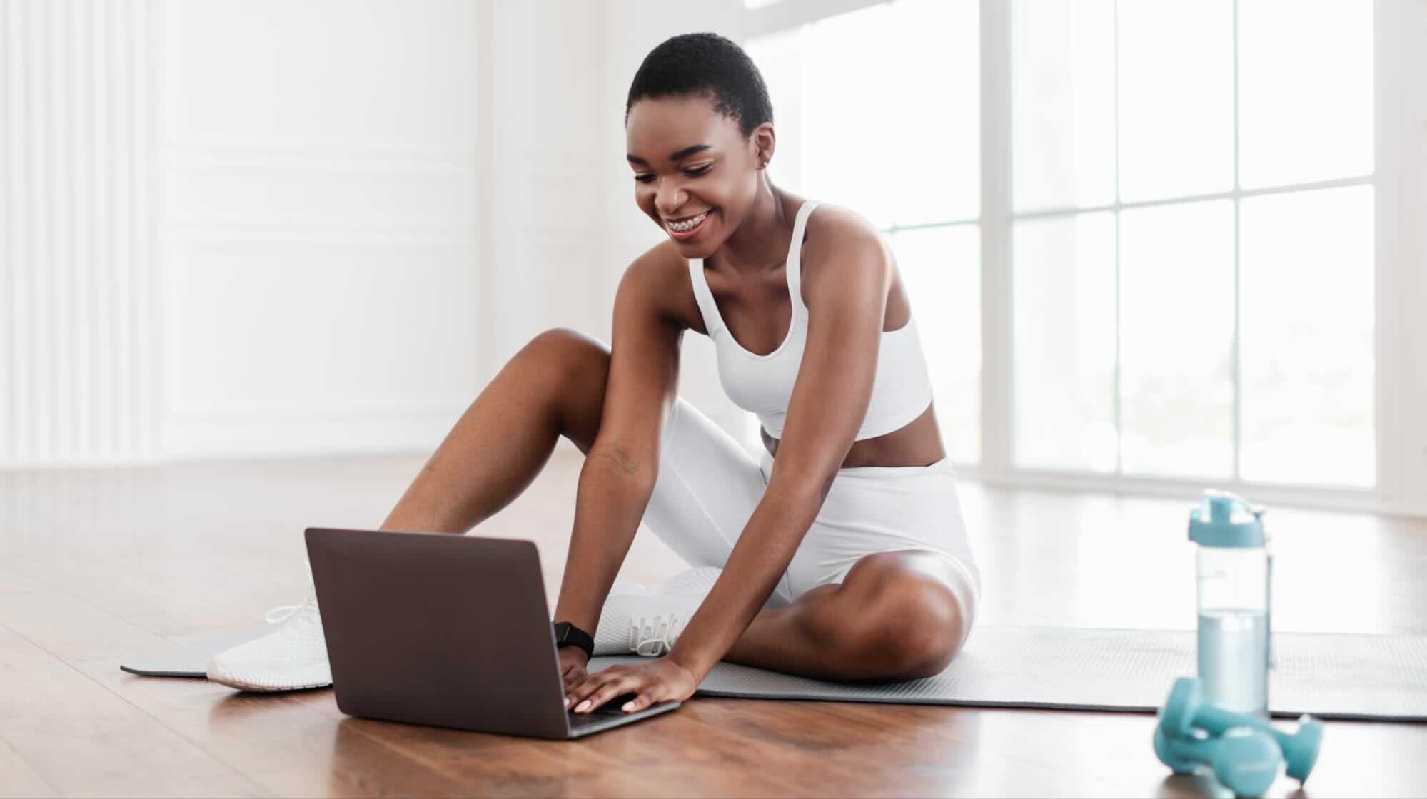 Mulher sorridente em roupa fitness branca sentada em um tapete de yoga usando um notebook, ao lado de uma garrafa de água e um halter, criando conteúdo para um blog de saúde e bem-estar.