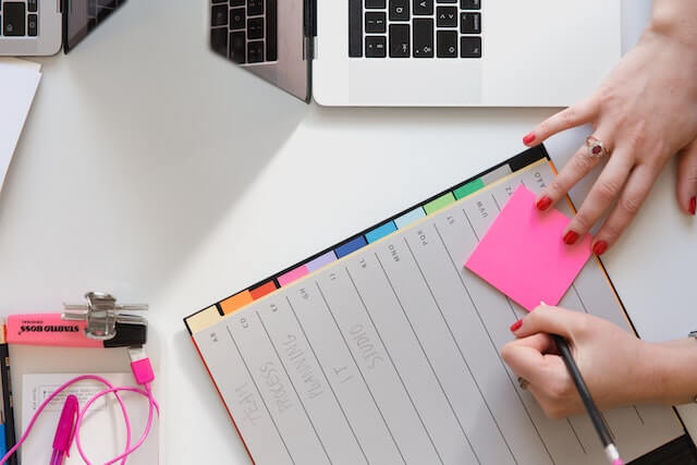 Person writing on pink sticky note on top of a planner