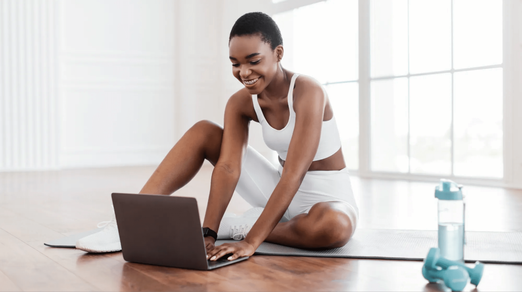 Mujer sonriente en ropa deportiva blanca sentada en una esterilla de yoga usando un portátil, junto a una botella de agua y una mancuerna, creando contenido para un blog de salud y bienestar.