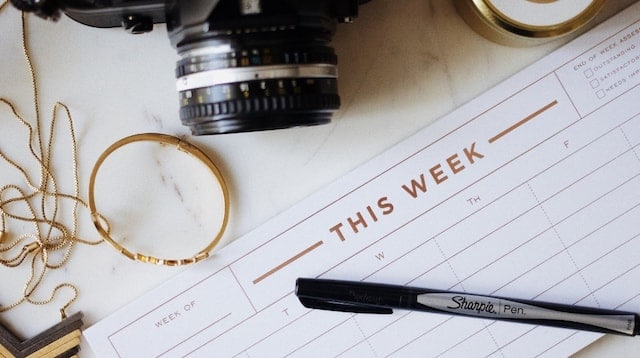 Agenda semanal sobre una mesa blanca junto a una cámara de fotos, un rotulador y accesorios dorados, representando la planificación editorial de un blog.