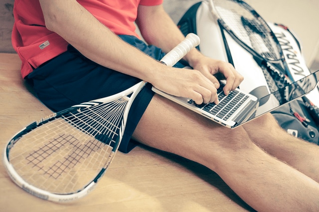 Persona en ropa deportiva sentada en el suelo escribiendo en un portátil junto a una raqueta y una mochila deportiva, creando contenido para un blog de salud y bienestar.