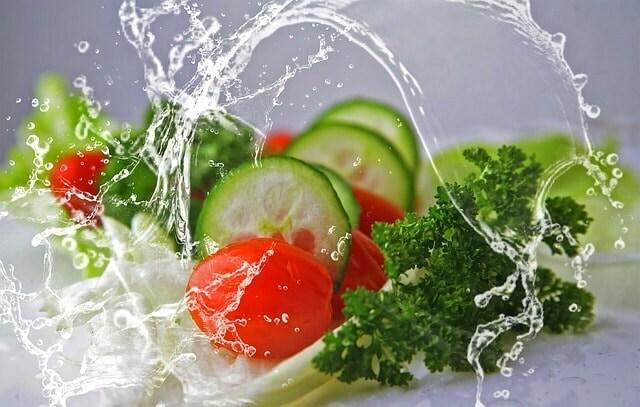 Tomates cherry, rodajas de pepino, lechuga y perejil frescos siendo bañados por un chorro de agua, ilustrando alimentación saludable y bienestar.
