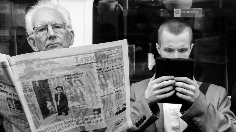 Man reading newspaper sitting next to man looking at tablet on the subway