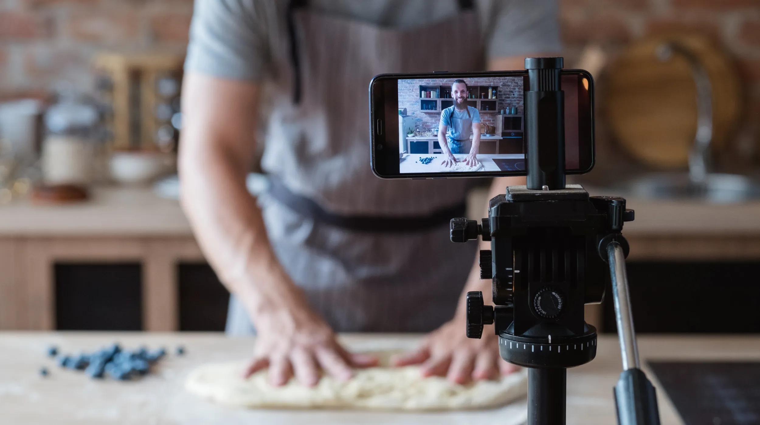 A person wearing an apron is filming themselves kneading dough in a kitchen, with a smartphone mounted on a tripod capturing the process.