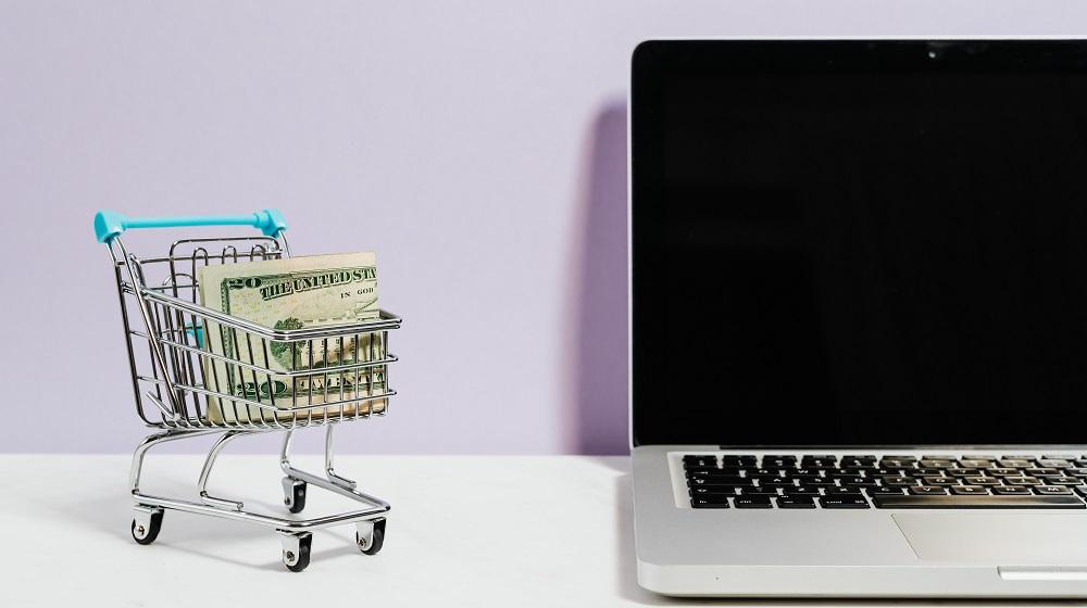 Tiny shopping cart holding a twenty-dollar bill near a laptop