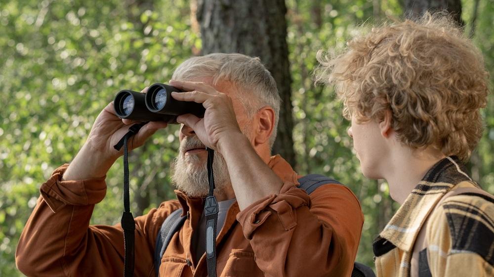 Two men in the woods with one looking through a pair of binoculars