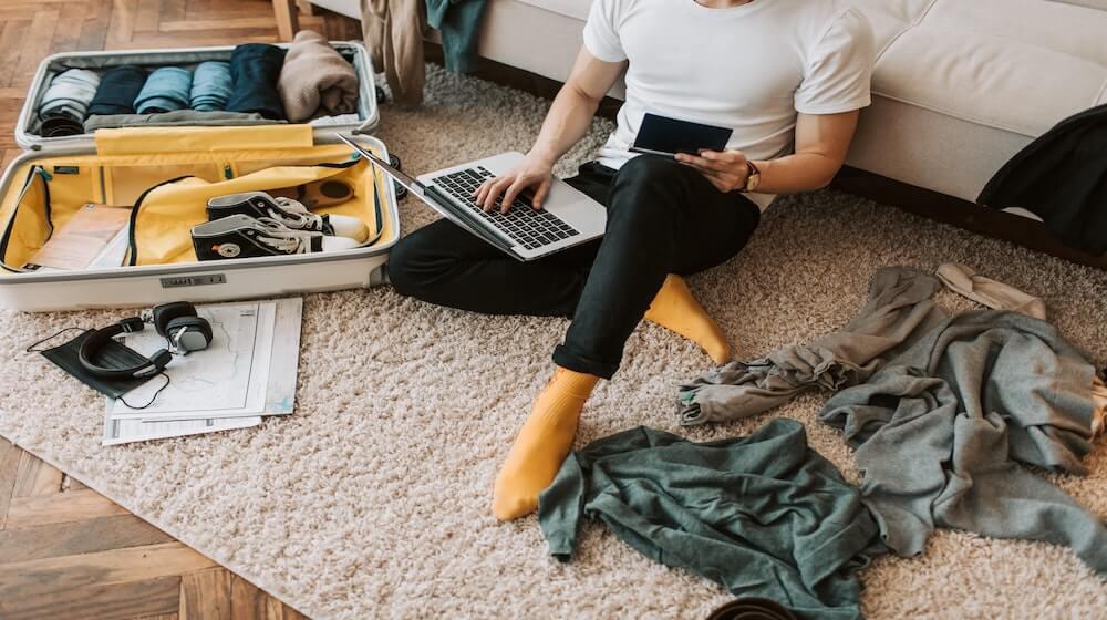 Man sitting on floor with laptop next to suitcase and scattered clothing items-2