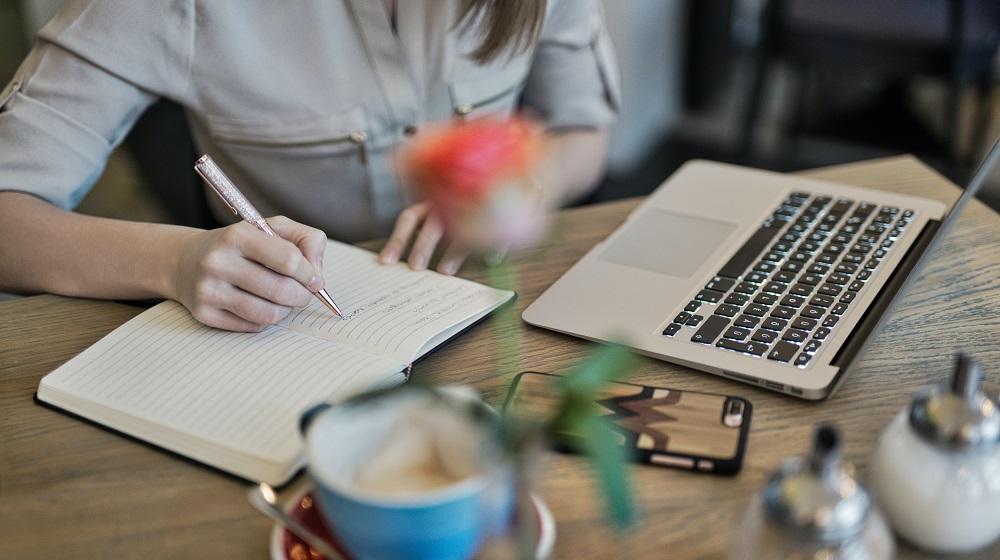 Woman writing in notebook with computer and phone nearby