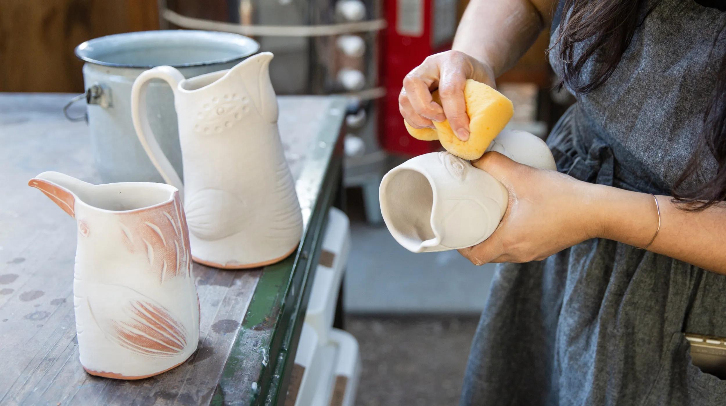 A person in a gray apron uses a yellow sponge to smooth the surface of a handmade ceramic pitcher shaped like a bird. Two other unique ceramic pitchers are on the table nearby, highlighting the type of handcrafted items often sold on Etsy vs eBay, where Etsy specializes in artisan and vintage goods, while eBay offers a wider range of products including mass-produced items.
