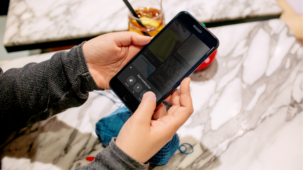 Person viewing Pinterest’s Idea Pin creation page on a smartphone over their crochet project that is resting on a stone table