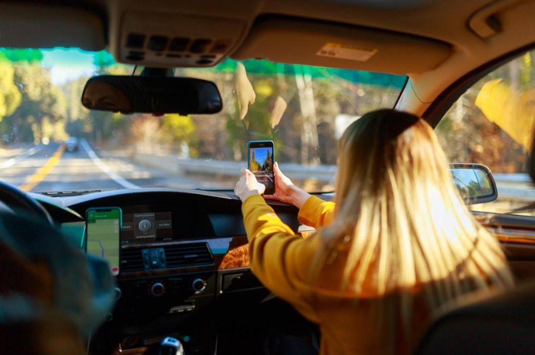 A woman facing away from the camera, taking a photo on her mobile phone in a car