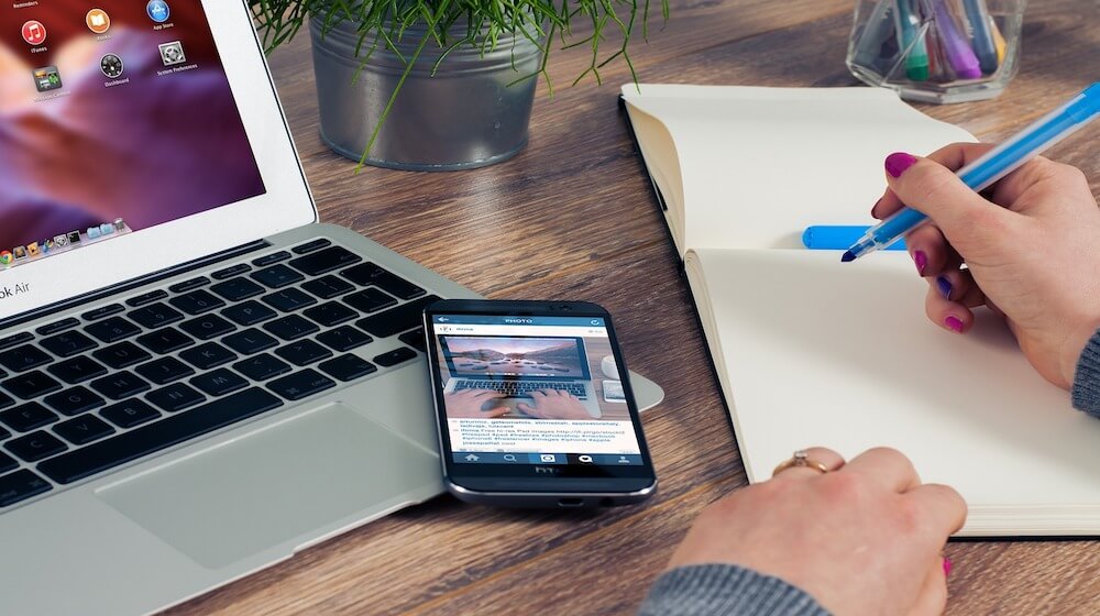 Close-up of person writing on notepad next to mobile phone and laptop, signifying how to write a business plan