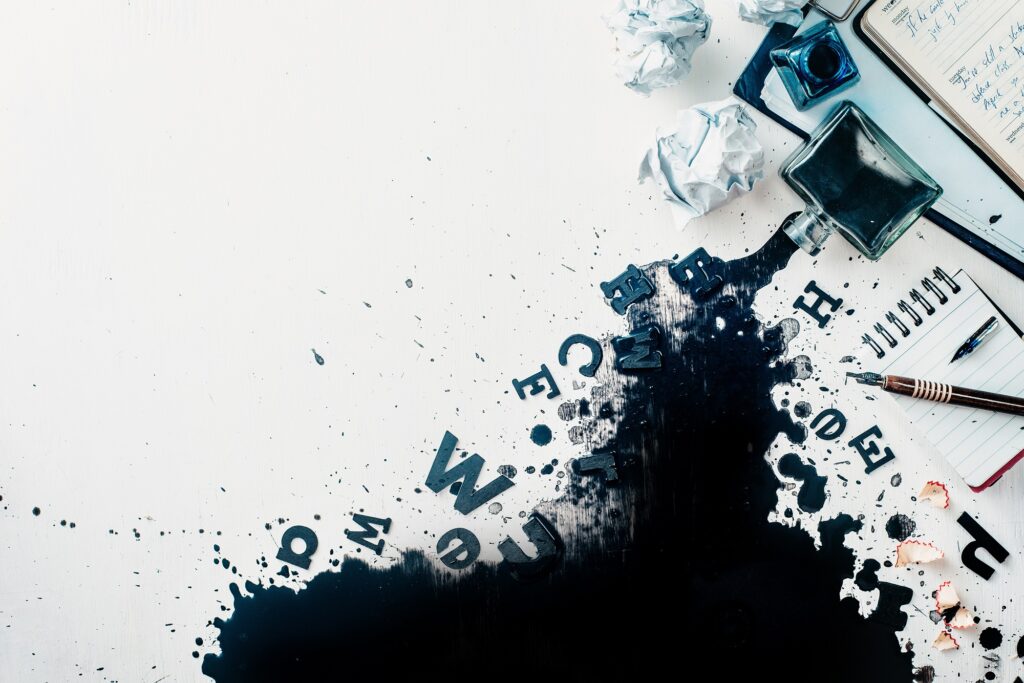 Header with spilled ink, crumpled paper, scattered letters, papers and notepads on a white wooden background. Creative writing concept. Flat lay with copy space. High key still life with an inkwell.