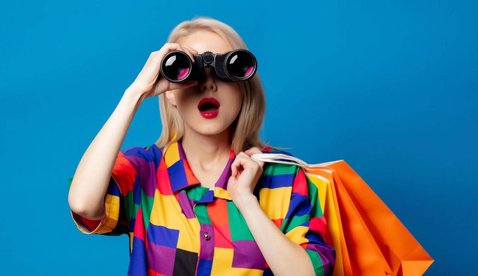 Woman in colorful outfit holding binoculers to face with mouth open in awe of something