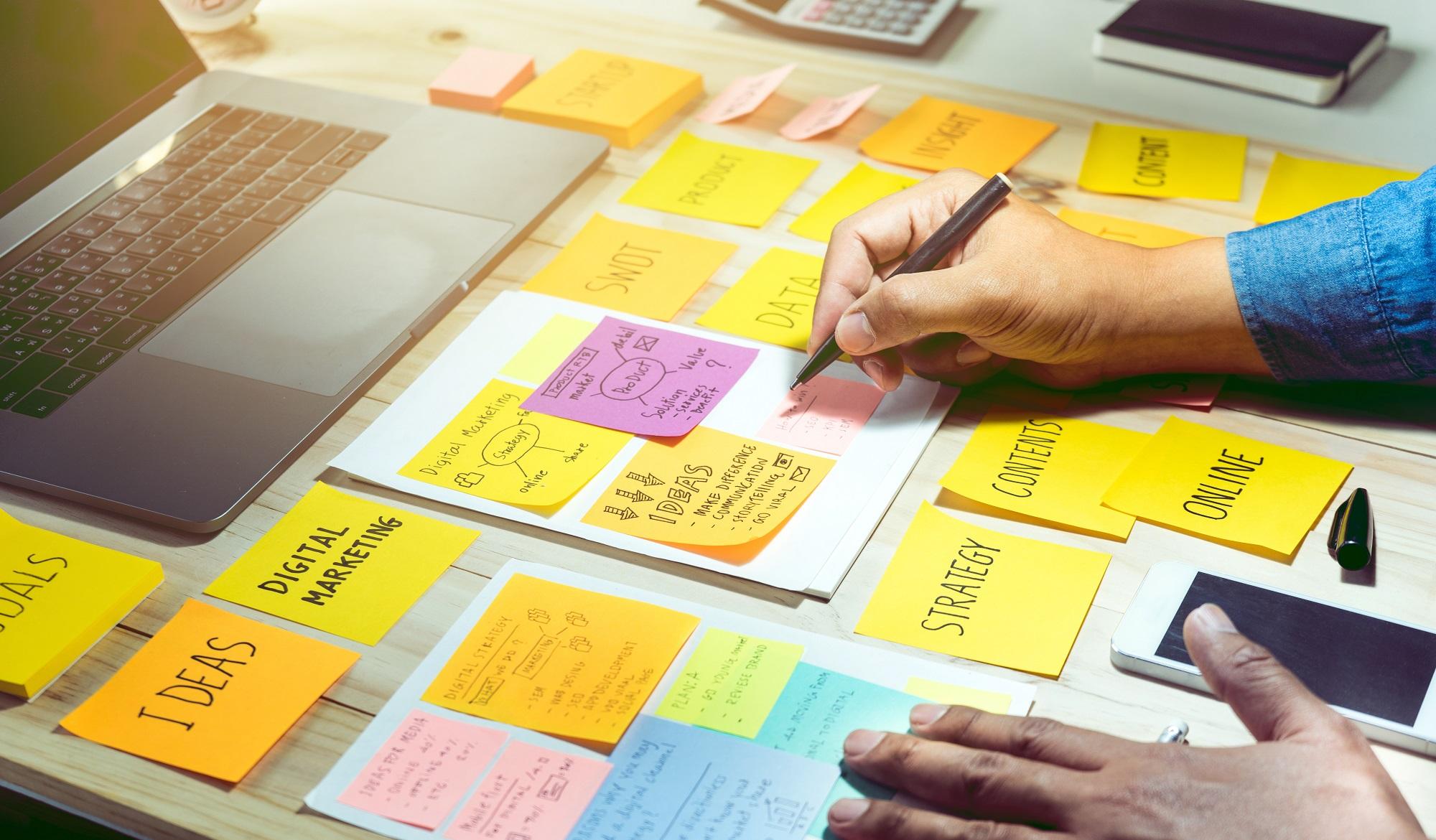 A HAnd Holding a Pen Is Poised over a Desk Covered in Sticky Notes