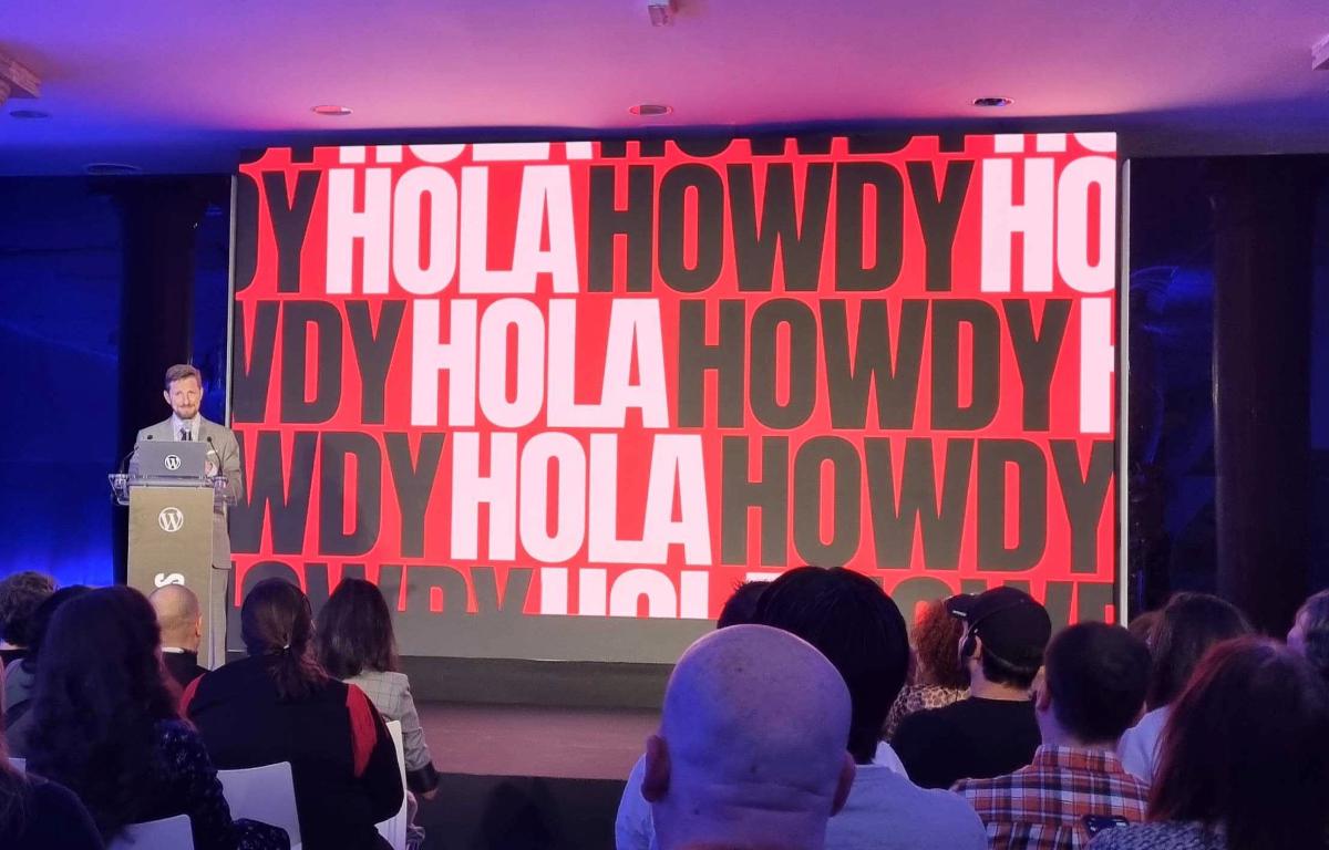 Matt Mullenweg stands behind a podium beside a screen reading Hola Howdy to the attendees at WordPress State of the Word 2023.