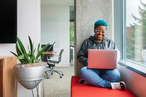 A joyful developer with blue hair works on open source WordPress projects on her laptop, seated comfortably on a red bench at GoDaddy's vibrant office space, reflecting the company's support for innovative and inclusive open source development