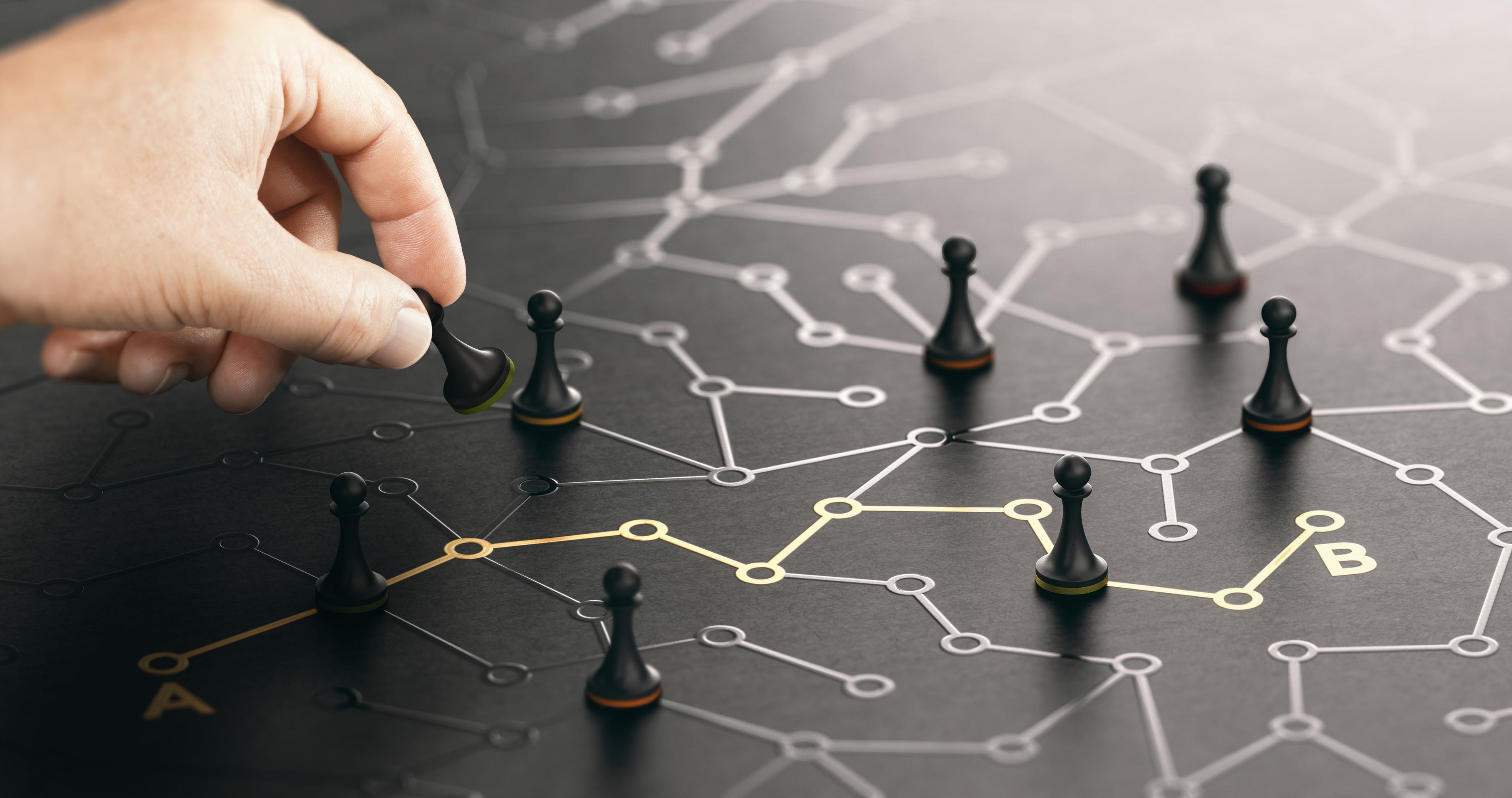 A hand moving a pawn on a conceptual board with other chess pieces, symbolizing strategic decision-making. The board features a network of lines and nodes with points labeled A and B, indicating a path or connection theme.