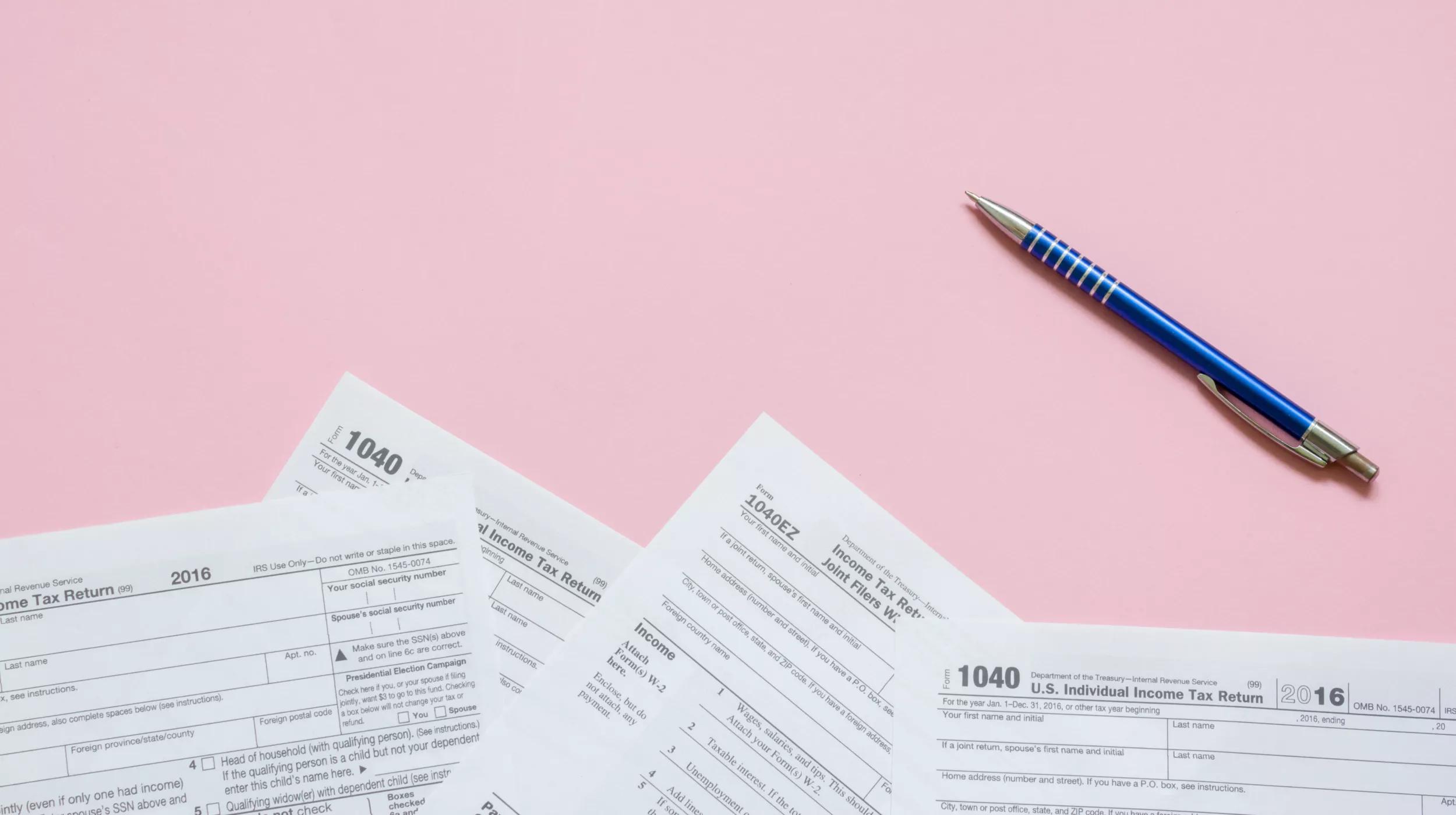 A flat lay of several U.S. tax forms, specifically the 1040 and 1040EZ forms for the year 2016, scattered on a soft pink background with a blue and silver pen resting diagonally across the top right corner.