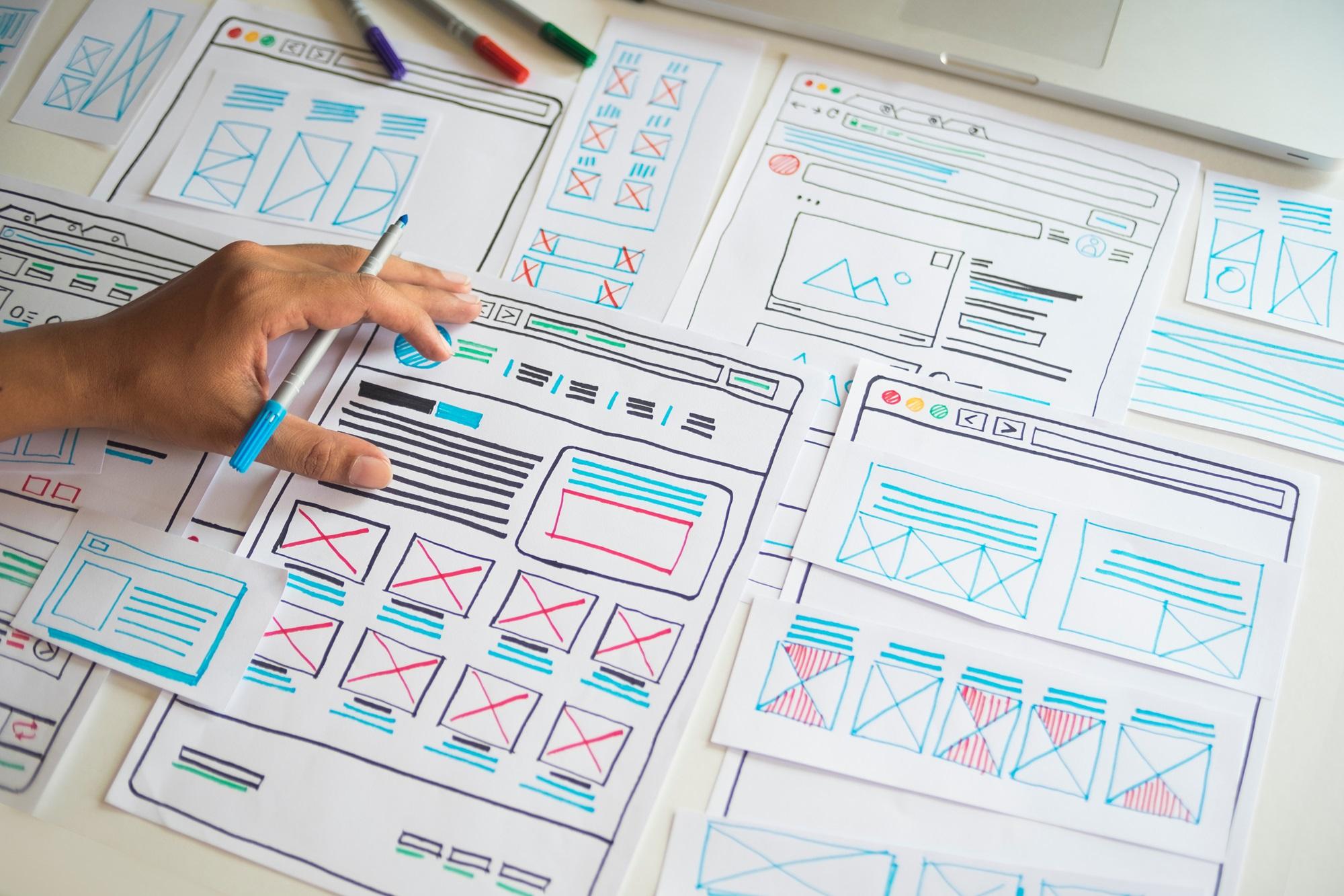 A person's hand holding a pen over a desk covered with various website wireframe sketches. The sketches include layouts for web pages with placeholders for images, text, and navigation elements. There are colored markers and a laptop partially visible in the background.