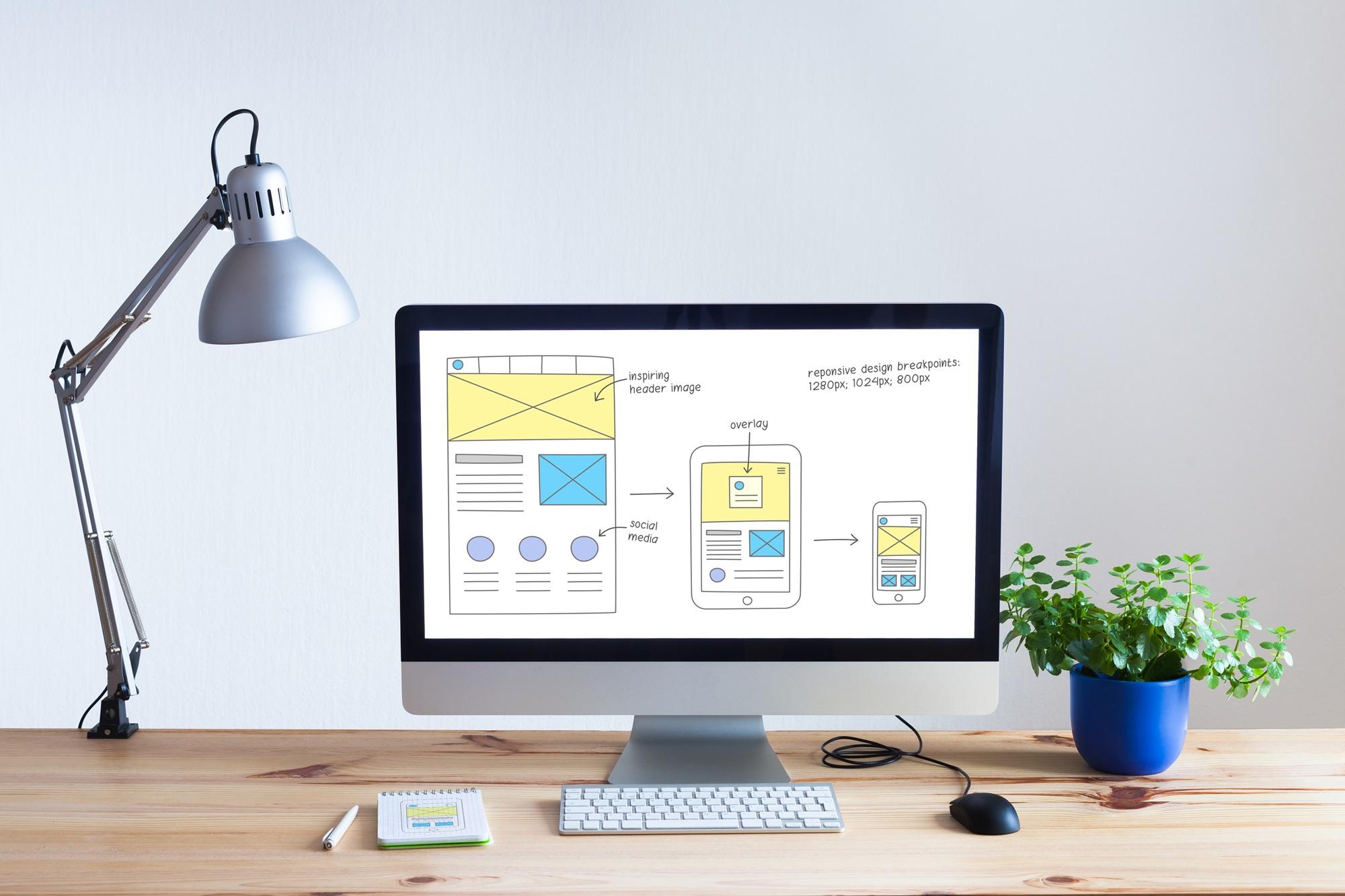 A clean and modern workspace with a desktop computer displaying a website design layout on the screen, a desk lamp to the left, a keyboard and a mouse in front, and a green potted plant to the right. The image on the screen includes a diagram illustrating responsive web design breakpoints for different device screens.