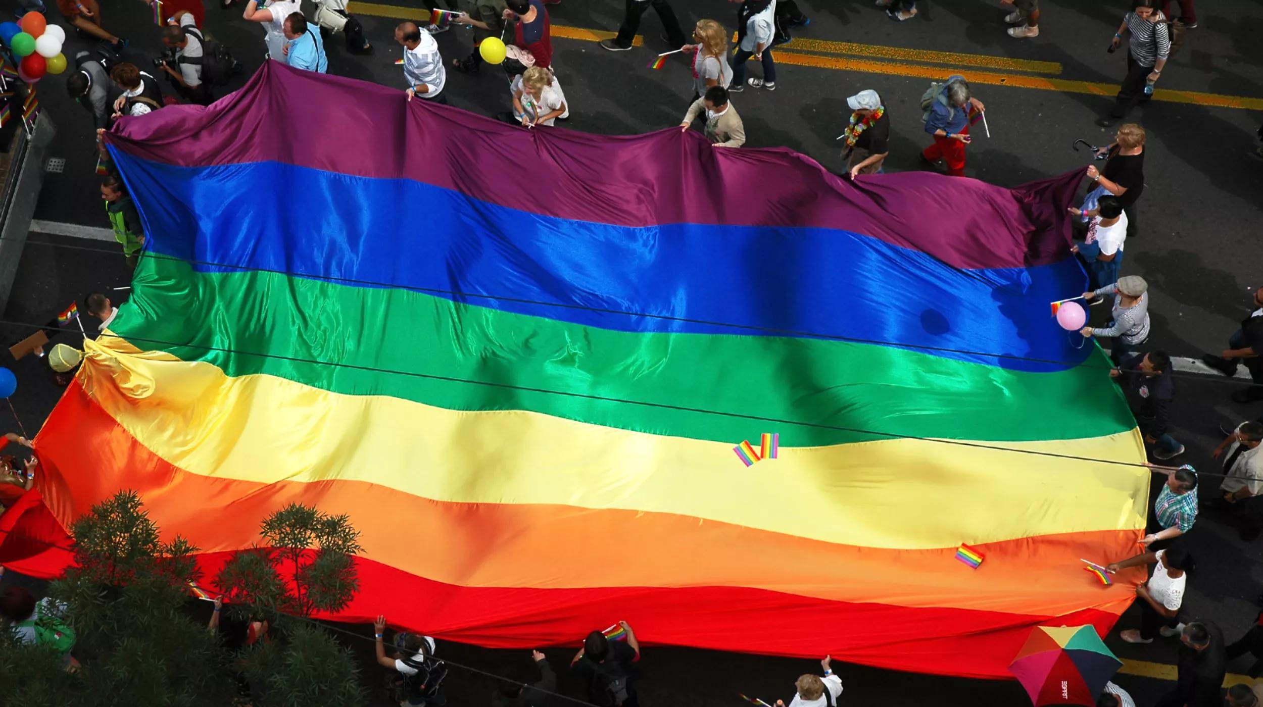 A large rainbow flag is carried by numerous people at a street parade. The flag, representing LGBTQ+ pride, extends across the width of the street, surrounded by participants, some of whom are holding smaller rainbow flags.