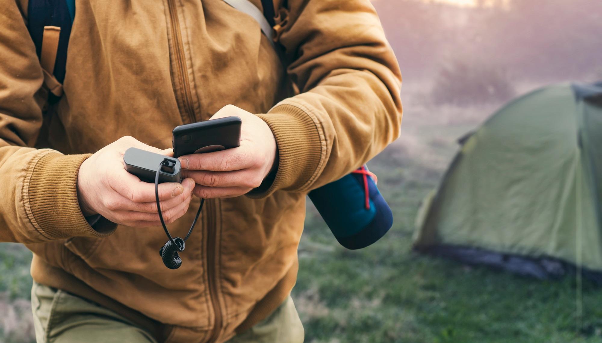 A person in outdoor clothing holding a portable power bank connected to a smartphone, with a green tent in the background, suggesting camping or outdoor activity.