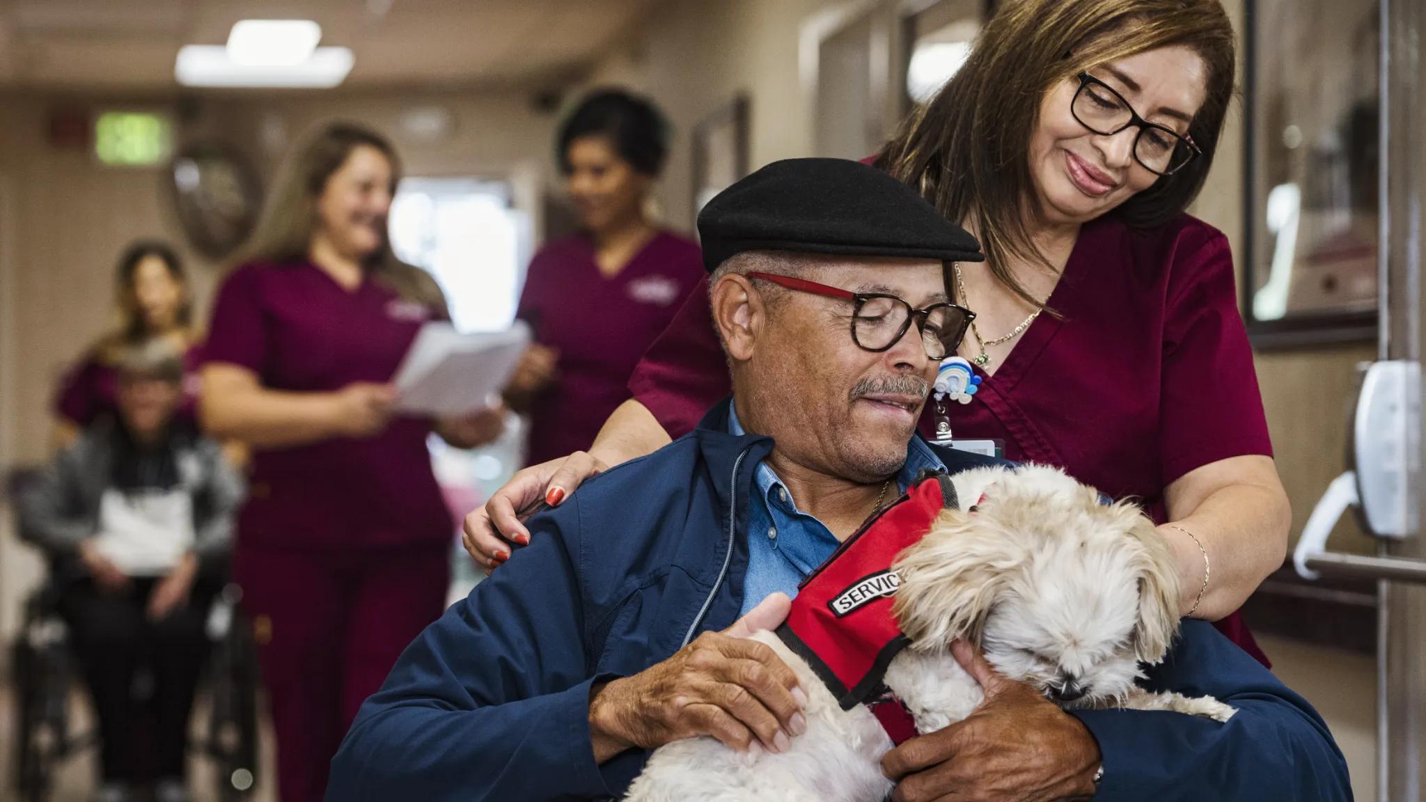 Healthcare worker watching over an elderly man holding a service dog.