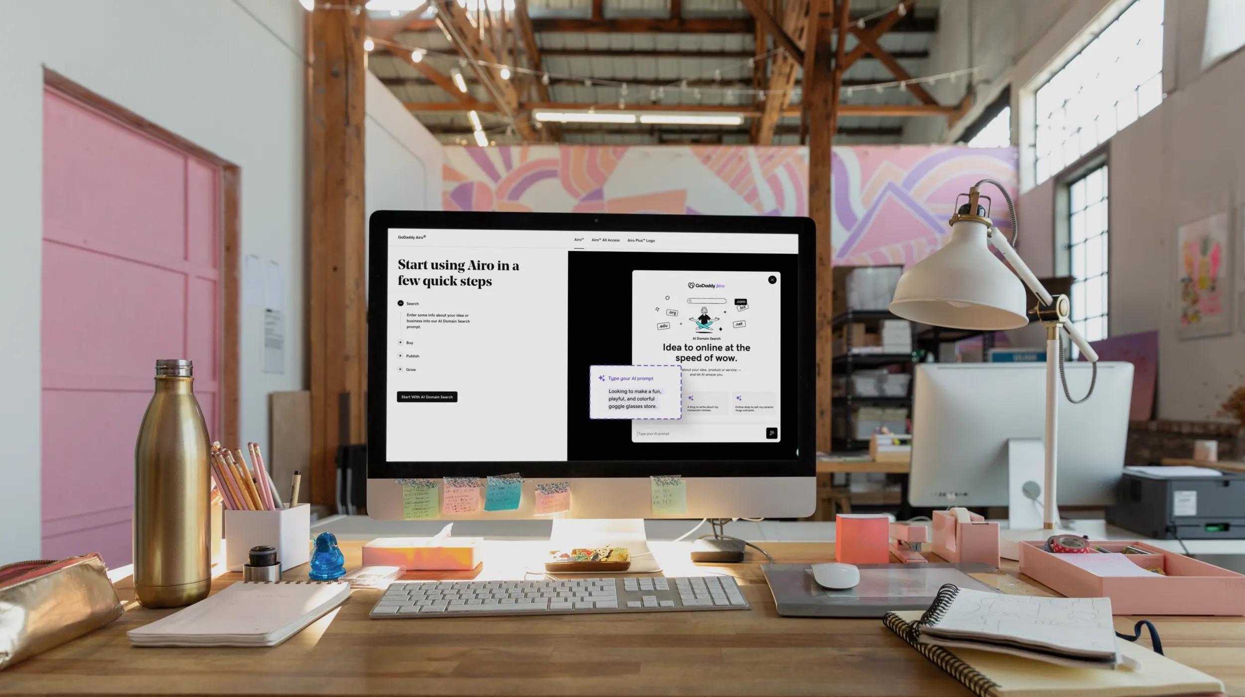 A modern, sunlit workspace with a desktop computer displaying the GoDaddy Airo setup screen. The desk is organized with a lamp, keyboard, notepad, water bottle, pencils, and sticky notes, creating a productive and creative office environment.