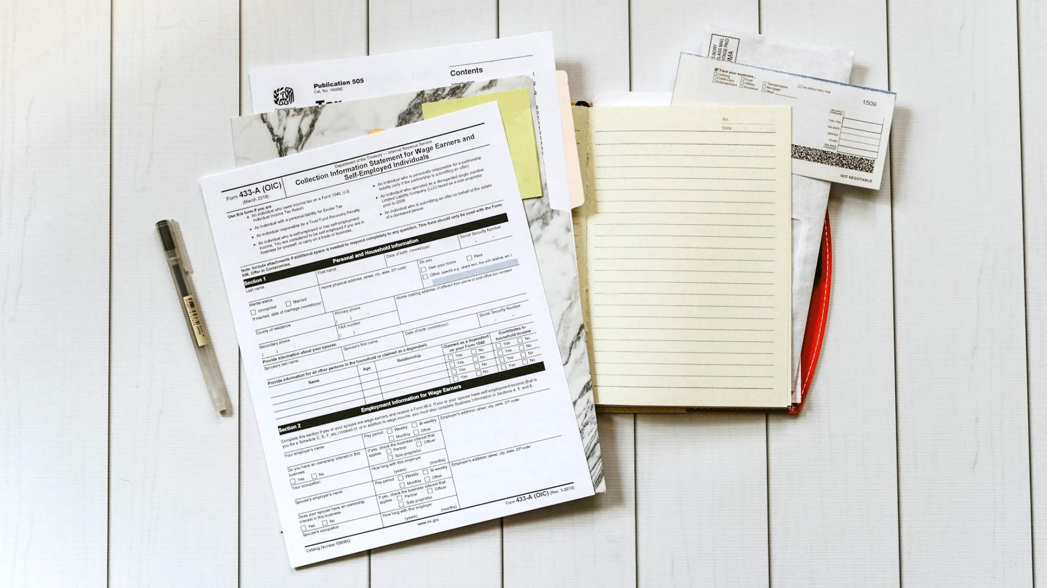 A collection of paperwork, including IRS tax forms, a pen, a lined notebook, and envelopes, arranged on a white wooden surface, illustrating the documents and steps needed to transfer an LLC to another state.