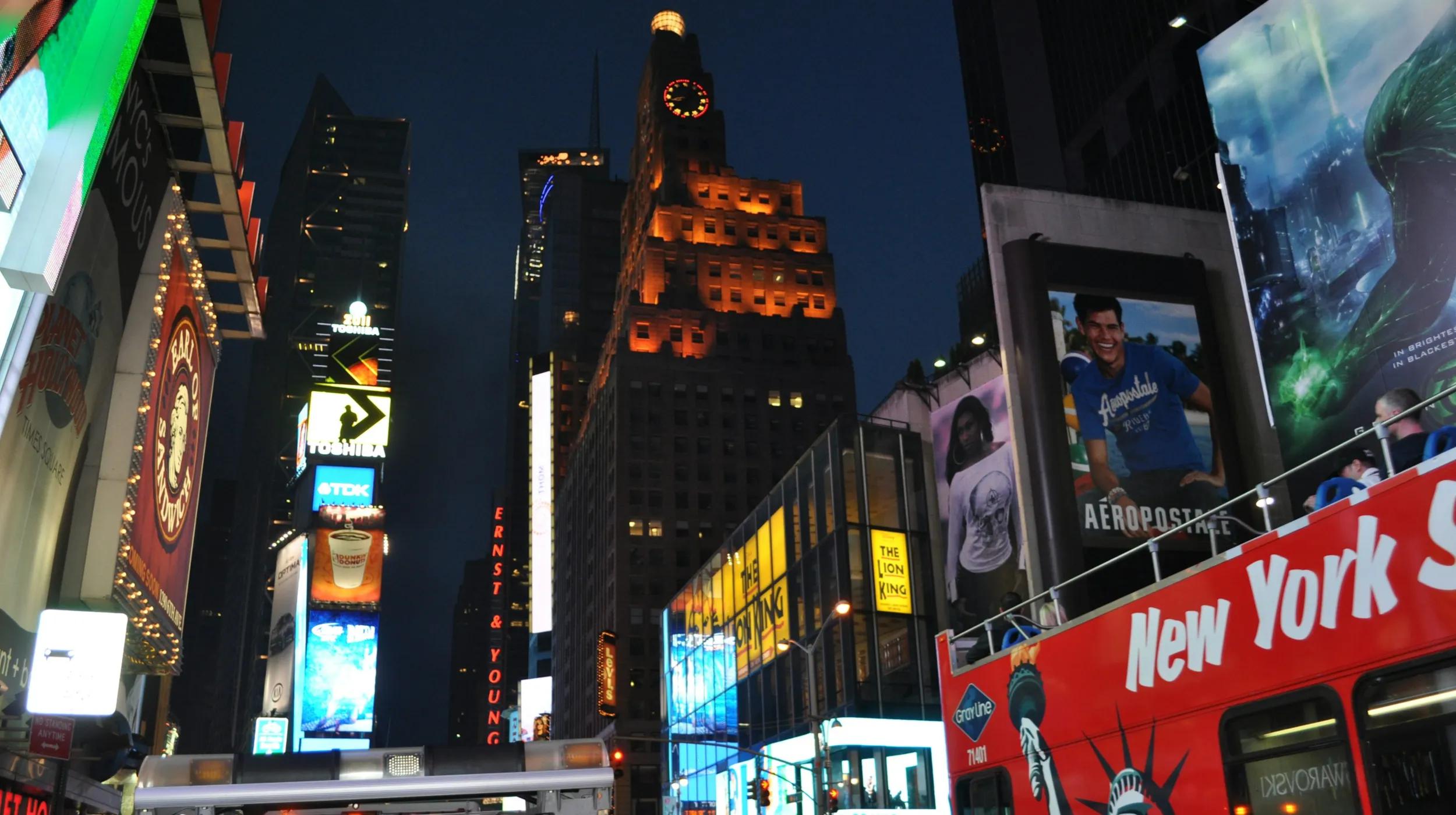 Alt text: Nighttime scene of Times Square in New York City, featuring brightly lit billboards, advertisements, and a red double-decker sightseeing bus with the words "New York" visible. Tall buildings and illuminated signs fill the background, with crowds and city activity partially visible.