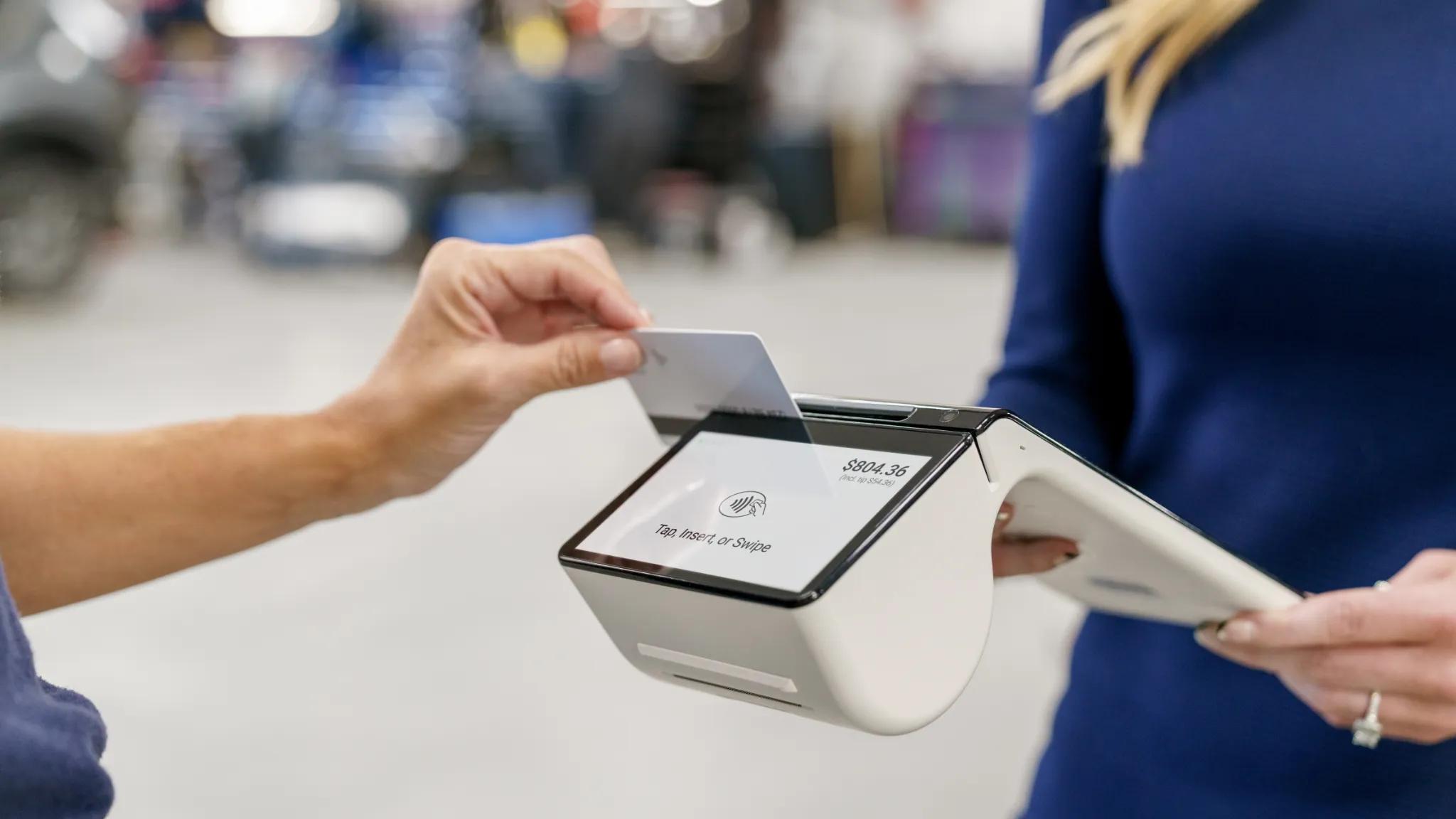 A customer swipes a credit card on a modern point of sale (POS) terminal held by a store employee; POS systems are used by businesses with a seller’s permit to process sales and collect required sales tax.