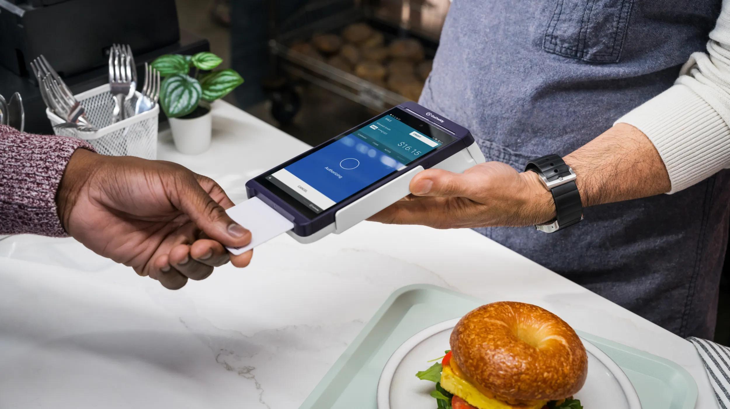 A customer inserts a credit card into a handheld retail POS system held by a worker in a café or restaurant, with a plate of food visible on the counter. The benefits of POS system technology are demonstrated here, making payments quick and convenient for both staff and customers.