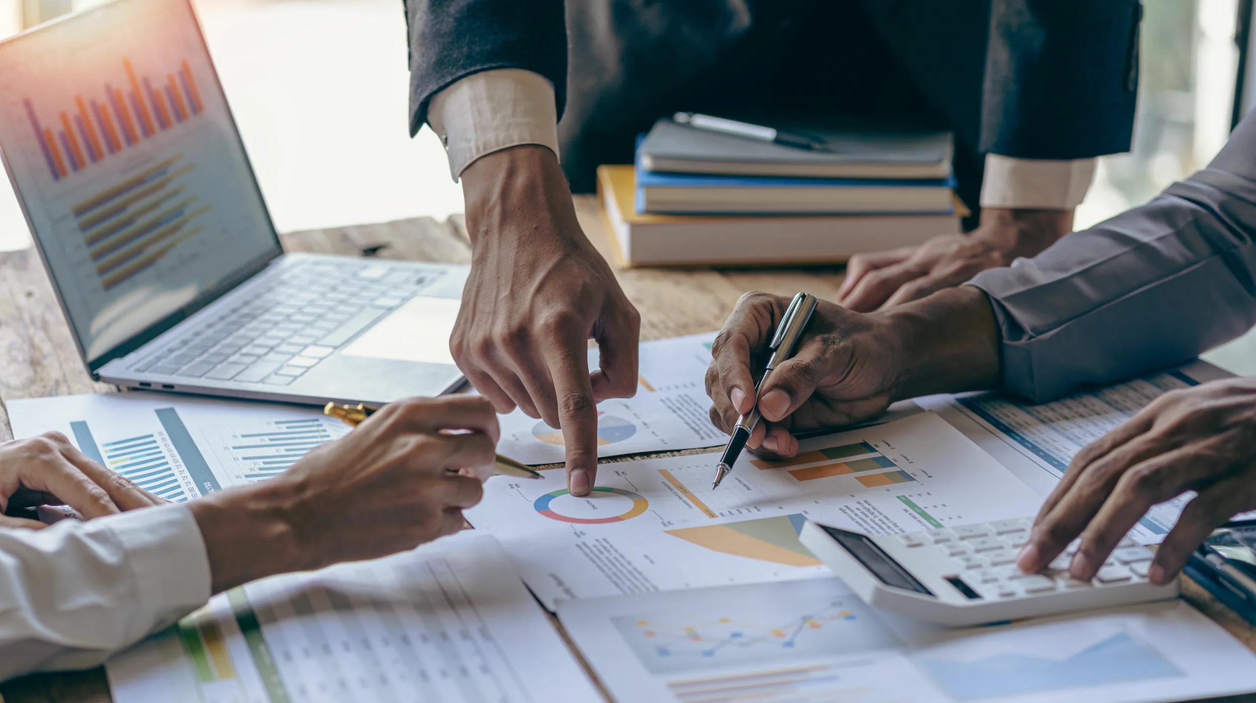 Three people review financial charts and graphs on paper with a laptop, books, and a calculator on the table, collaborating on data analysis and planning for business operations.