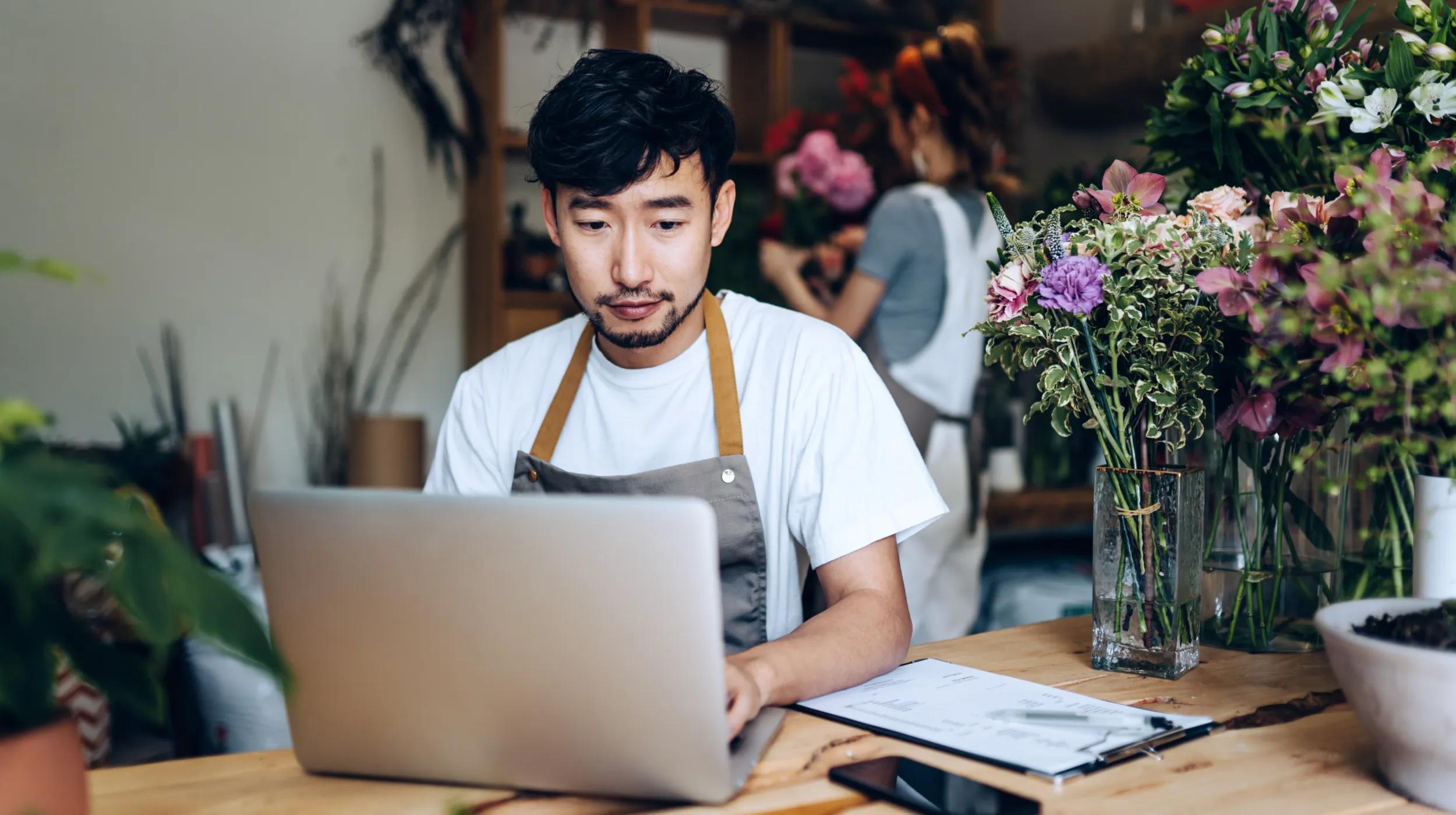 A florist wearing an apron sits at a wooden table surrounded by flowers, working on a laptop with a clipboard nearby, appearing focused as he looks up how to write a business proposal for his flower shop.