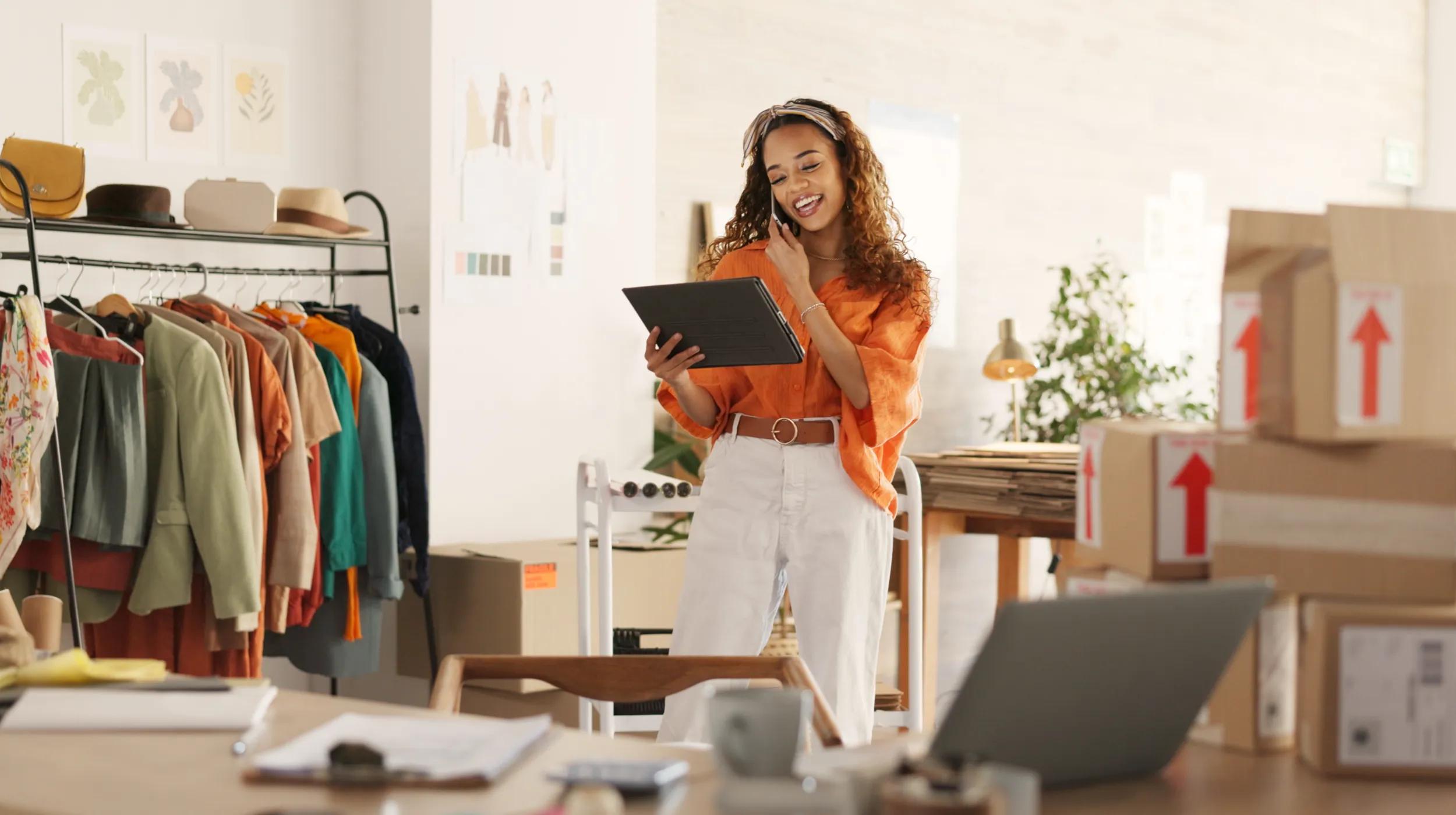 A woman in an orange shirt stands in her boutique, smiling as she talks on the phone and uses a tablet to generate ai products descriptions for her clothing items. Clothing racks, shipping boxes, and a laptop are visible around her workspace.