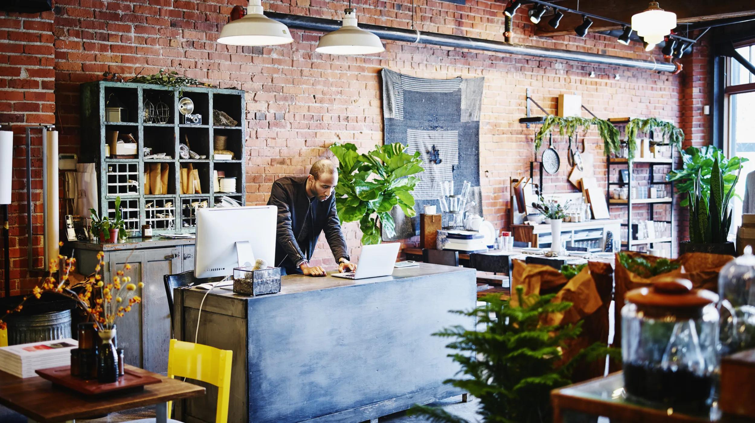 A small business owner stands behind the counter of a stylish shop with exposed brick walls and lots of plants, reviewing documents on a laptop, possibly working on an operating agreement for the business.