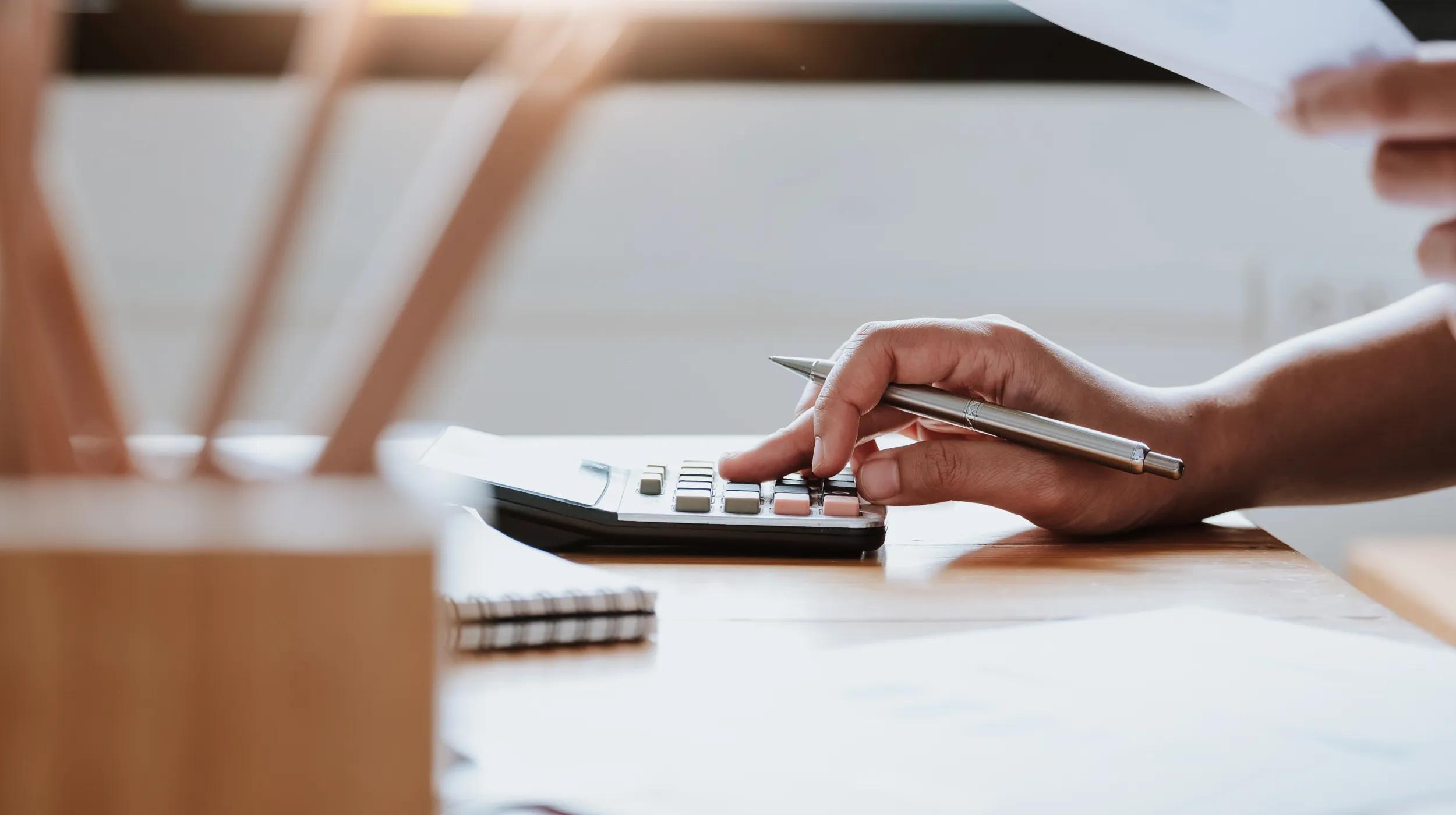 A person using a calculator and holding a pen at a desk, illustrating how to calculate sales tax for financial or business purposes.