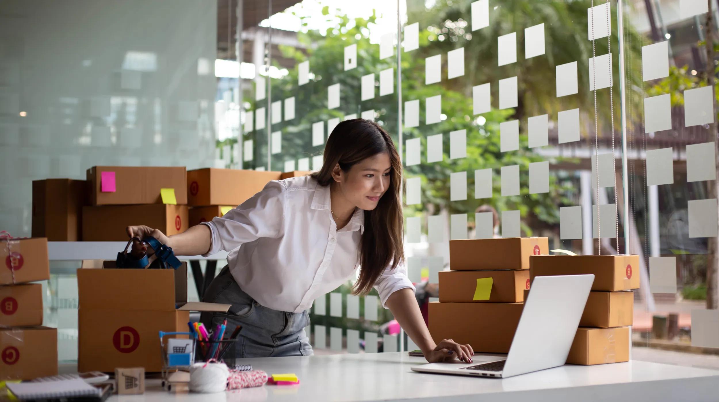 A young woman in a white shirt stands at a desk in a modern office, surrounded by stacks of cardboard boxes and looking at her laptop, representing the organization and shipping tasks involved in running a subscription business model naturally. The workspace is bright with large windows and sticky notes on boxes, suggesting an efficient small business operation.